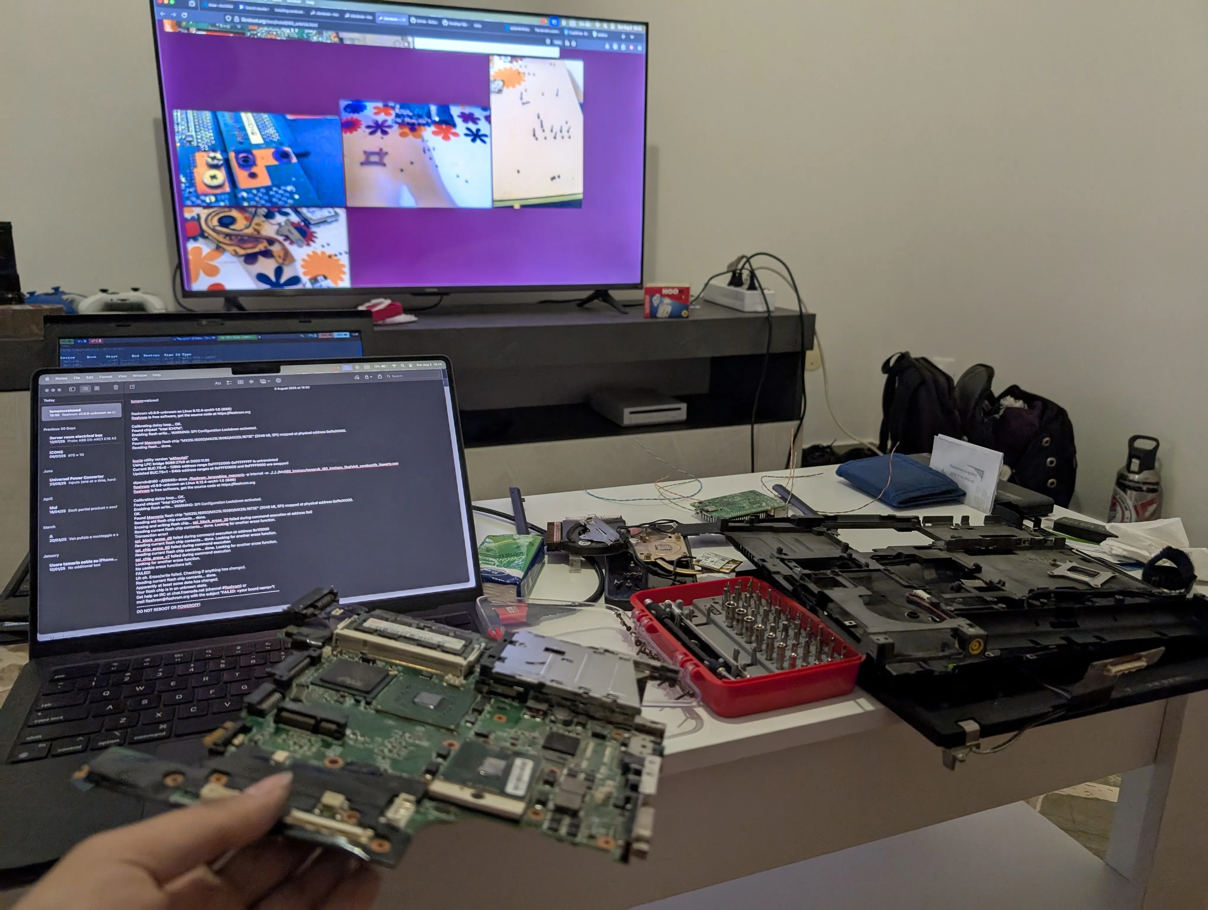 The ThinkPad T60 motherboard is held in the foreground, while, in the background, there&#x27;s a little table holding a MacBook with our notes displayed, the rest of the T60 chassis and display, a
screwdriver kit, and other bits and pieces. In the far view, a television is showing a webpage with photos of other disassembled
T60&#x27;s.